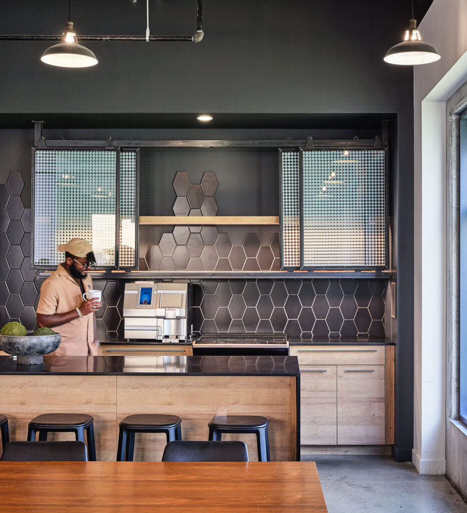A man wearing a cap and mask stands in a modern kitchen, holding a cup near a coffee machine. The kitchen features black hexagonal tile backsplash, wooden cabinets, and bar stools along a counter.