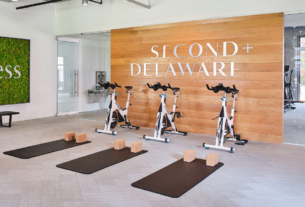 A modern fitness room features three stationary bikes and three yoga mats with blocks, set on a light floor. The wall behind has wood paneling with “SECOND + DELAWARE” in large white letters. A green plant wall is on the left.