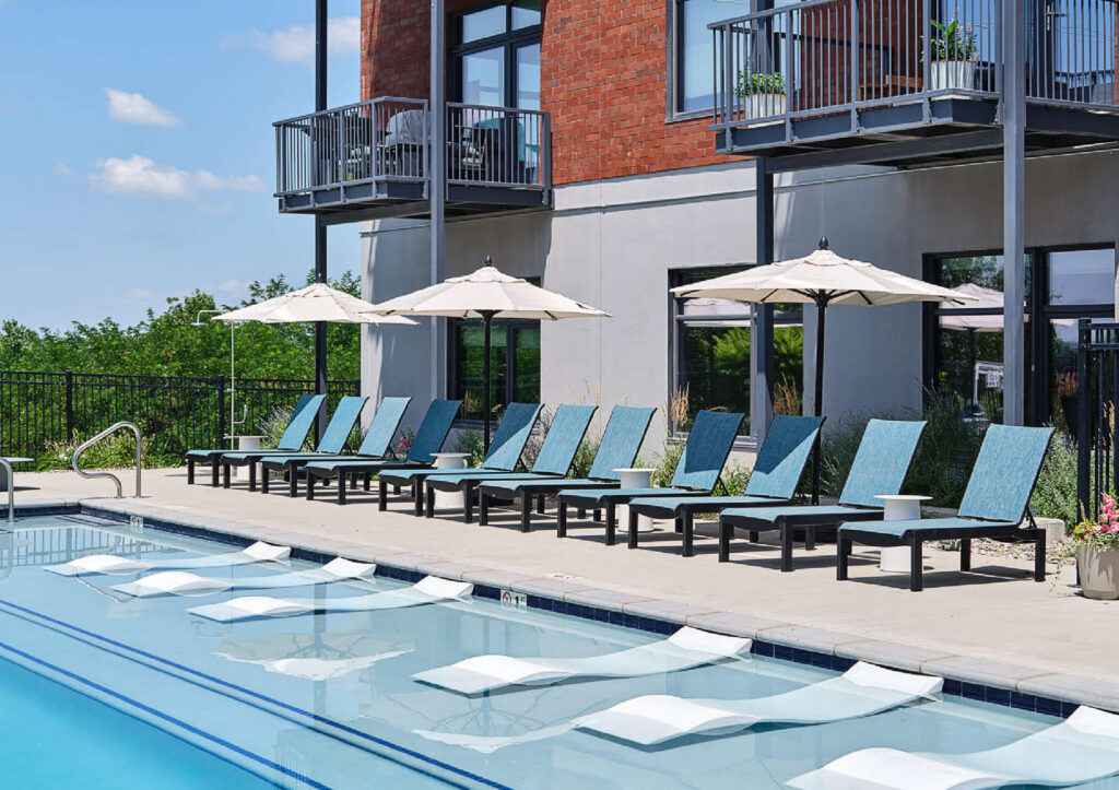 Outdoor swimming pool with lounge chairs and umbrellas lined up along the deck. Some loungers are partially submerged in the shallow pool area. Modern apartment balconies overlook the scene. Trees and blue sky in the background.