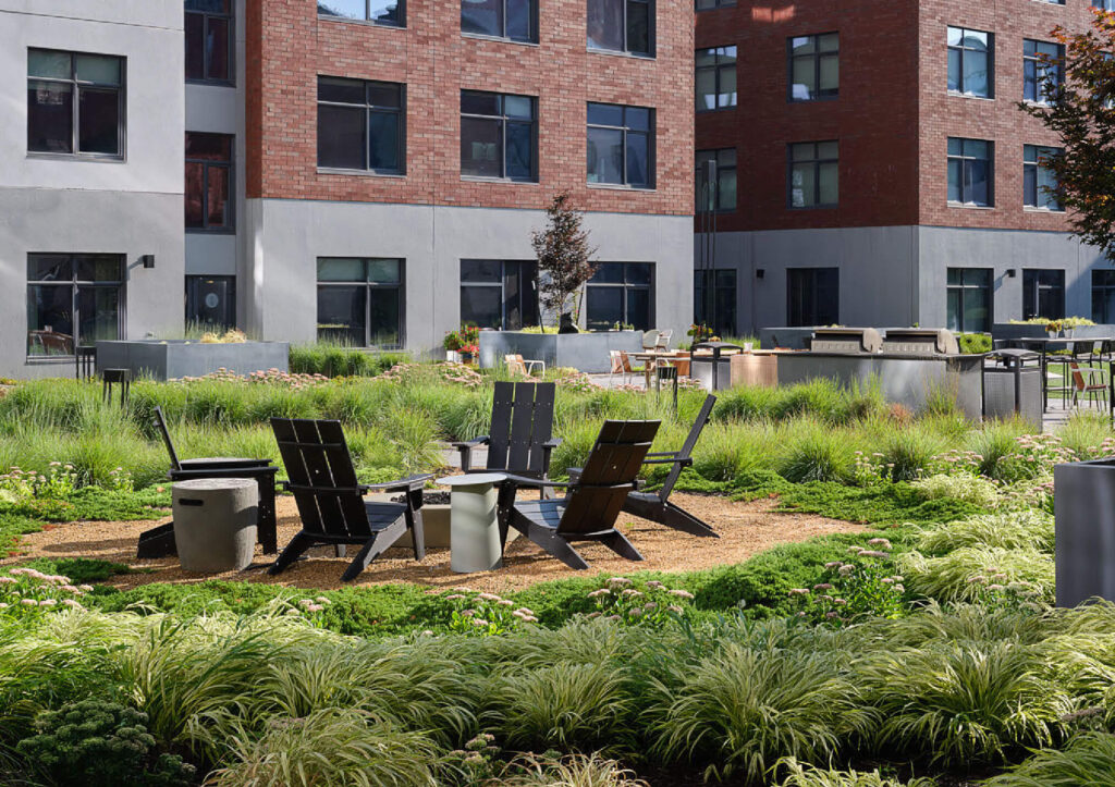 A modern apartment courtyard with Adirondack chairs arranged around a fire pit, surrounded by lush green plants and grasses, outdoor tables, and a grill area near brick and gray buildings.