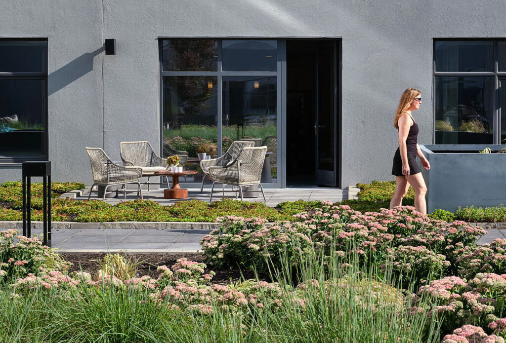 A woman in a black dress walks past blooming plants in a garden, with a modern building and outdoor seating area featuring four chairs and a table in the background.