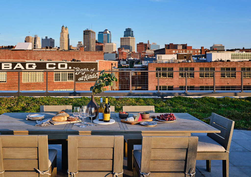 A rooftop patio with a wooden dining table set for a meal, including wine, bread, grapes, and tableware, overlooking a city skyline with brick buildings and high-rise towers at sunset.