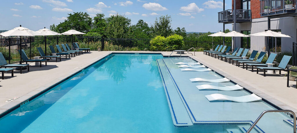 Rectangular outdoor swimming pool with lounge chairs, poolside umbrellas, and several built-in loungers partially submerged in the water. Trees and a building are visible in the background under a blue sky.