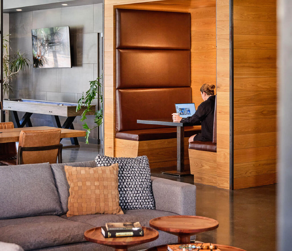 A person works on a laptop in a brown leather booth set into a wood-paneled wall. In the foreground is a gray couch with textured pillows and round wooden coffee tables. A pool table and plants are in the background.