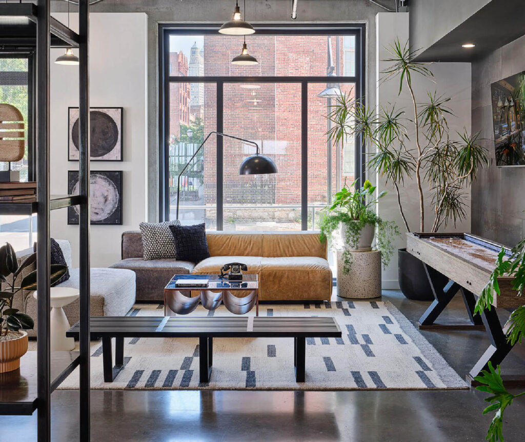 Modern living room with a brown and white sectional sofa, patterned rug, potted plants, large windows, wall art, a floor lamp, and a shuffleboard table, featuring an industrial and cozy design.