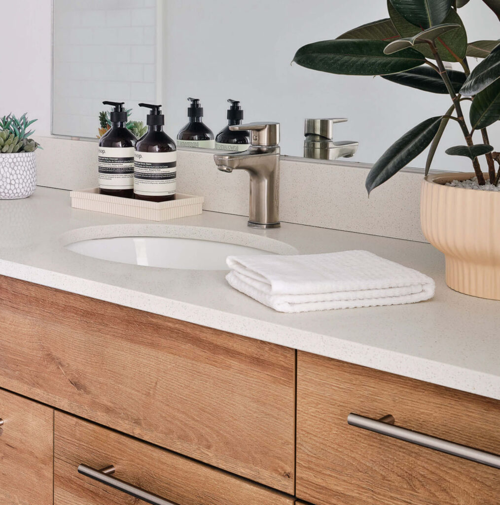 A modern bathroom vanity with a white countertop, wood drawers, a silver faucet, neatly folded white towels, liquid soap bottles, small potted plants, and a large mirror in the background.