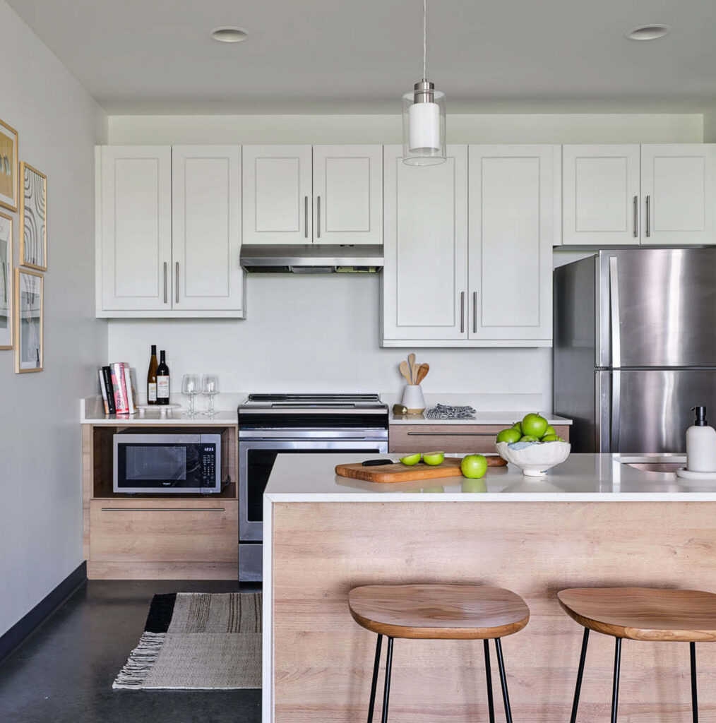 Modern kitchen with white cabinets, stainless steel appliances, a microwave, and a kitchen island. Two wooden stools are by the island, which holds a bowl of green apples, a soap dispenser, and a wooden tray.