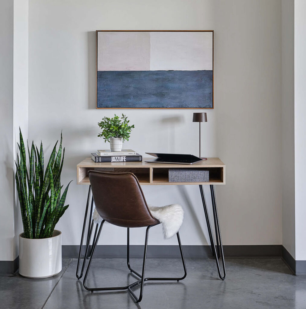 A modern minimalist workspace with a wooden desk, brown leather chair with a white cushion, small lamp, plant, books, and a framed abstract painting above. A tall potted snake plant stands beside the desk.