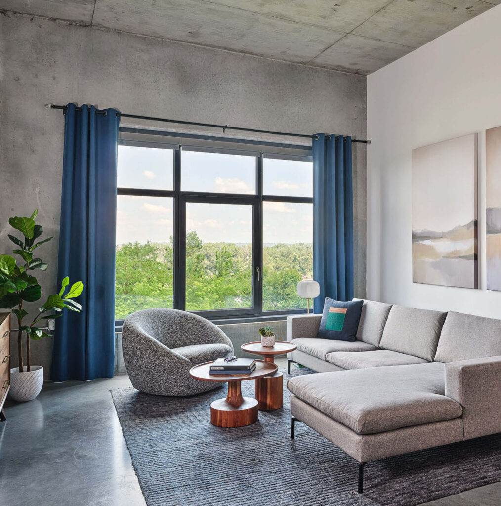A modern bedroom with a wooden bed, white bedding, a black-and-beige rug, potted plants, a brown armchair, wooden dresser, large window with beige curtains, and abstract artwork on a gray wall.