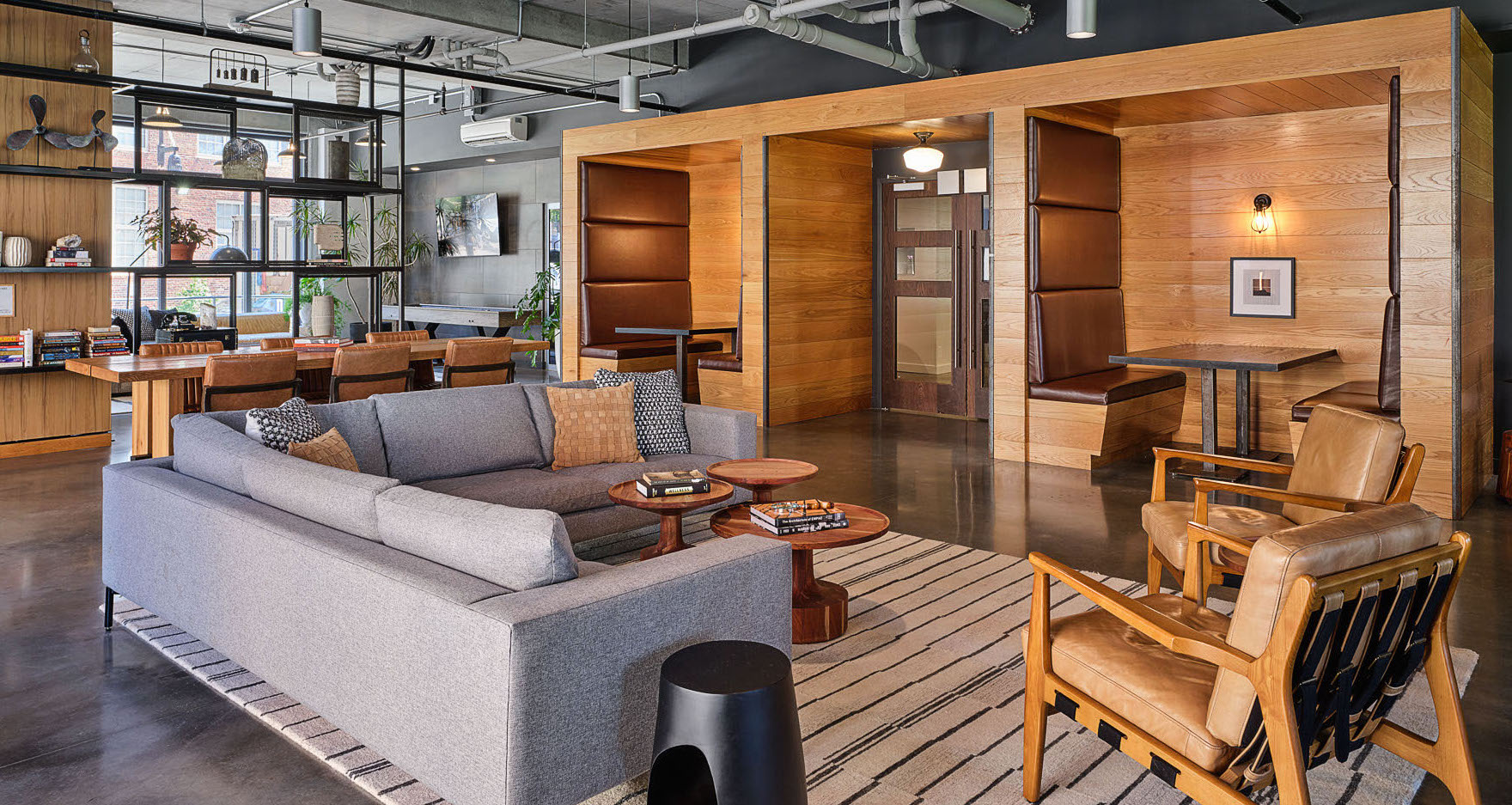 Modern office lounge with a gray sofa, wooden chairs, and a striped rug. In the background, private booth seating is built into a wooden alcove. Open shelves and desks line the back wall, with large windows providing natural light.