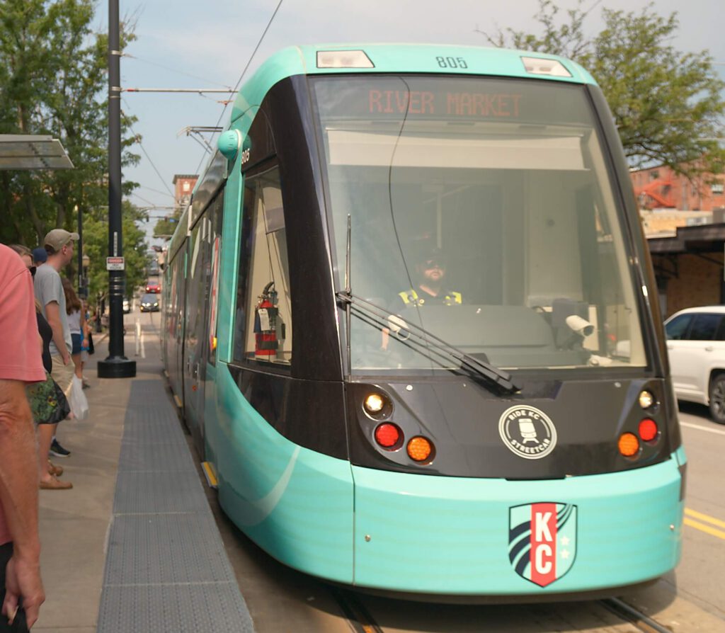 A teal KC streetcar labeled River Market stops at a sidewalk in the neighborhood. A person stands nearby, with trees and buildings lining the street in the background.