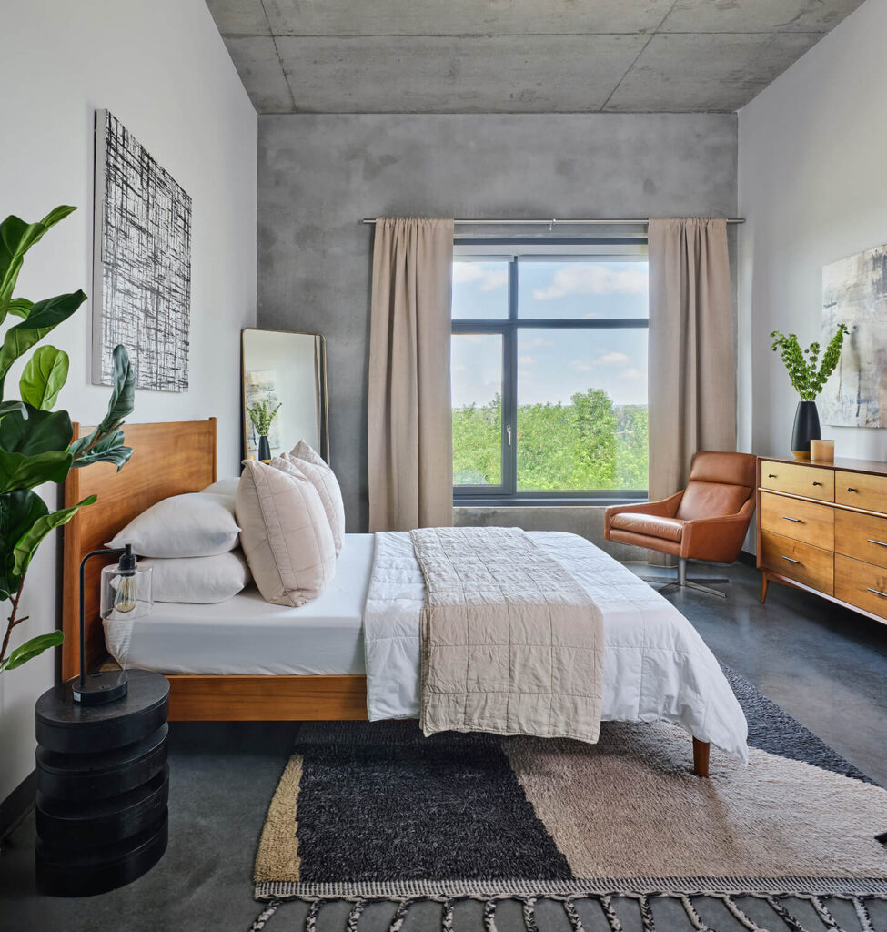 A modern bedroom with a wooden bed, white bedding, a black-and-beige rug, potted plants, a brown armchair, wooden dresser, large window with beige curtains, and abstract artwork on a gray wall.