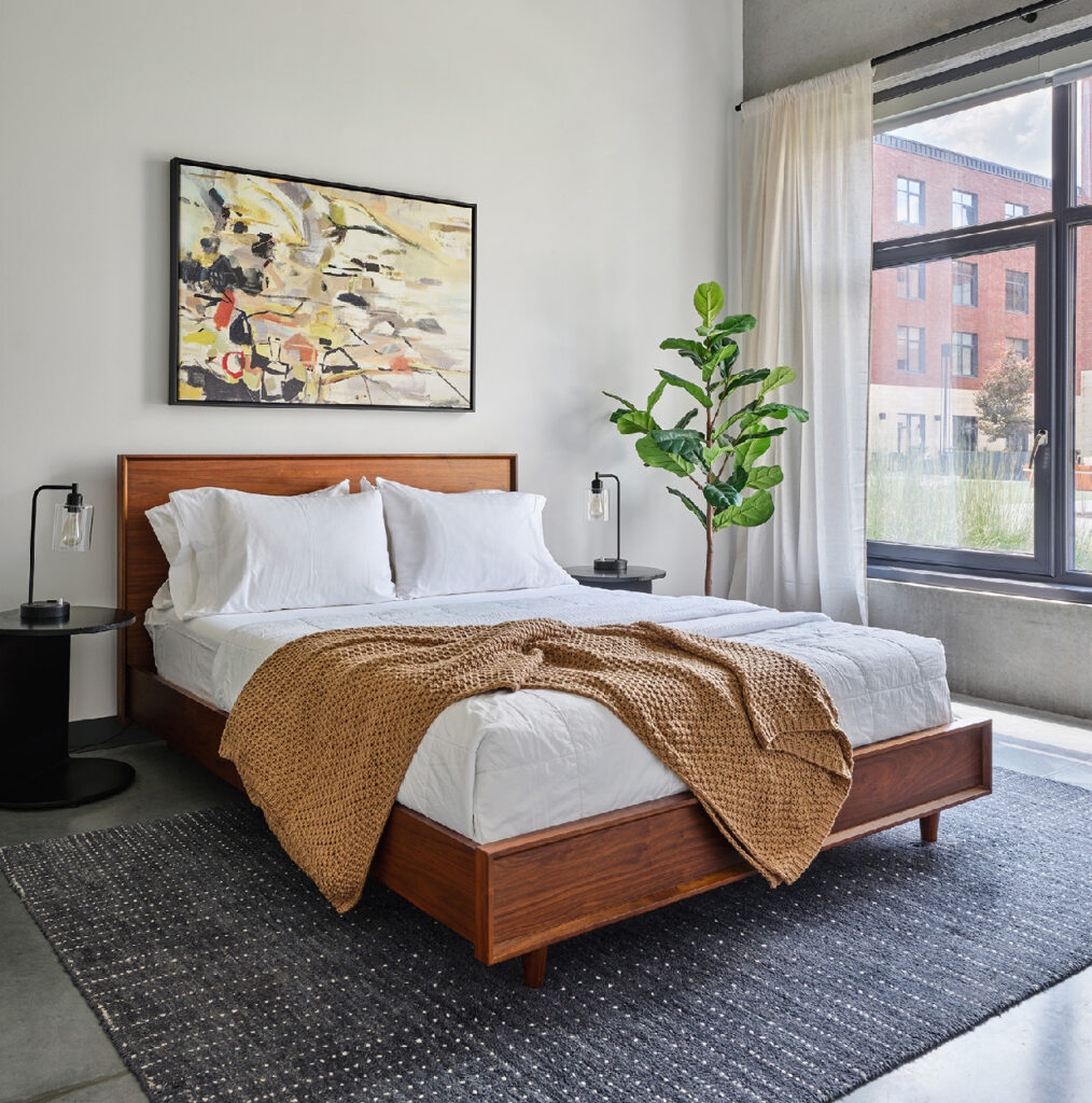 A modern bedroom features a wooden bed with white bedding and a tan blanket, flanked by black nightstands and lamps. A large plant and abstract painting add decor, while sunlight streams in through a large window with white curtains.