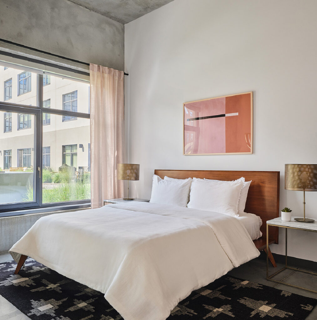 A modern bedroom with a wooden bed frame, white bedding, two gold lamps on side tables, abstract art on the wall, pink curtains, and large windows overlooking an apartment building courtyard.