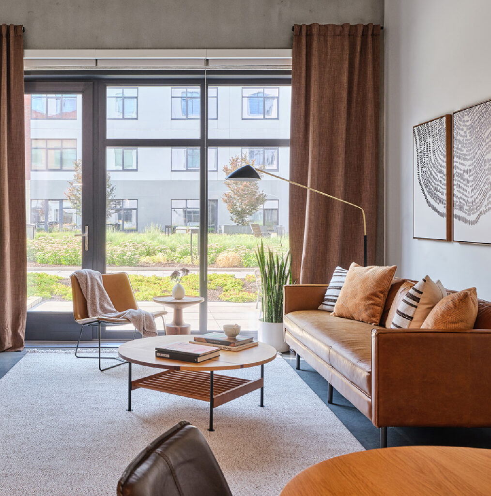 Modern living room with a tan leather sofa, striped pillows, round coffee table, floor lamp, armchair, large window with brown curtains, and a view of greenery and apartment buildings outside.